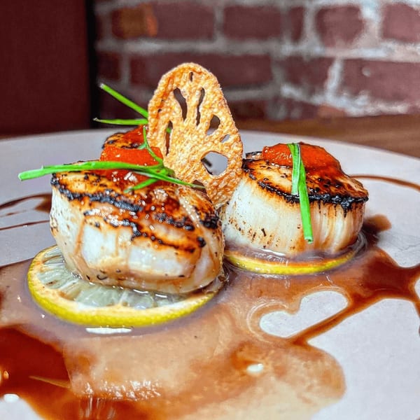 Seared scallops on lemon slices with red sauce, chives, and a lotus root chip on a white plate.