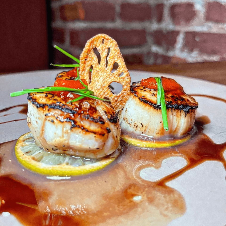 Seared scallops on lemon slices with red sauce, chives, and a lotus root chip on a white plate.